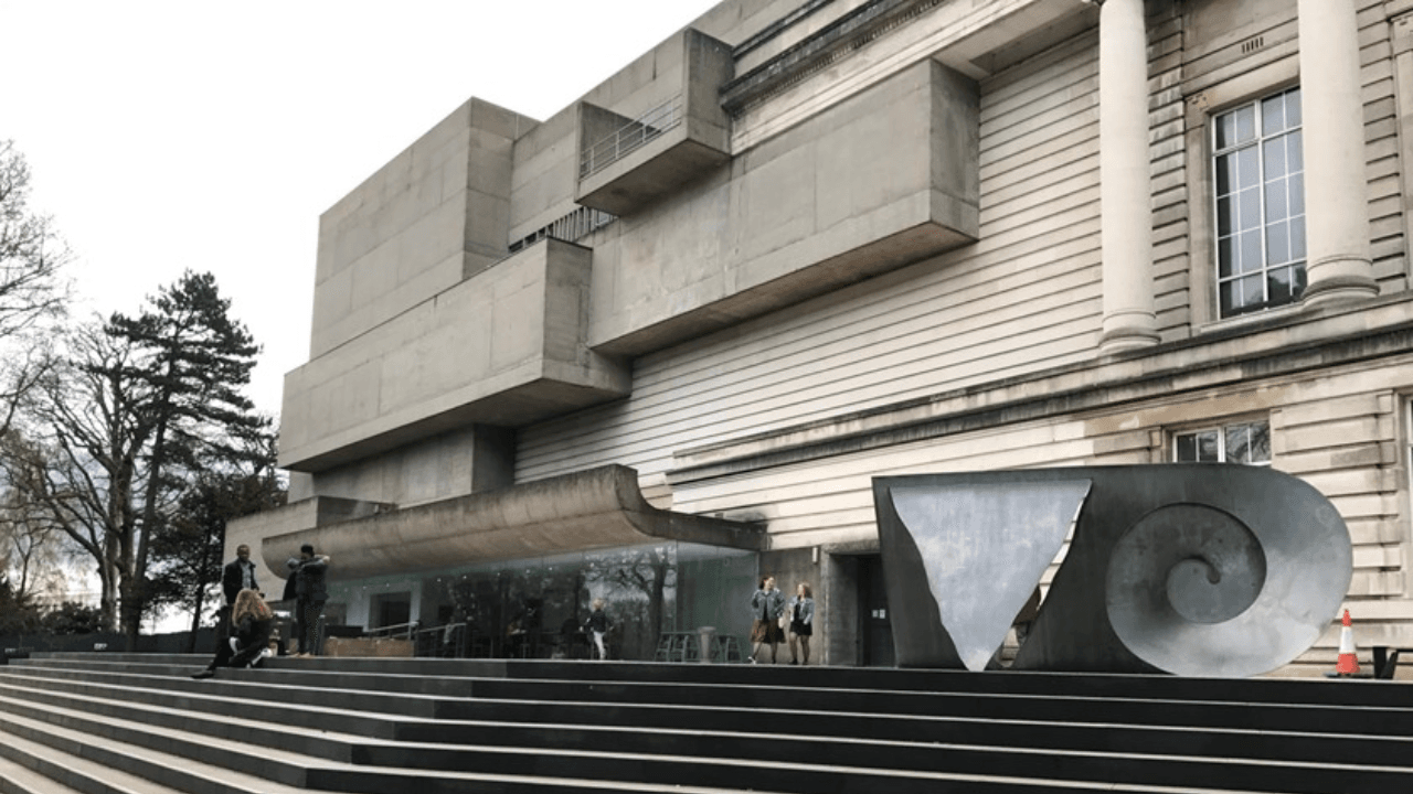 The front of the Ulster Museum, a modernist building with a mix of stone and concrete surfaces, large windows, and a prominent entrance set within a landscaped area.